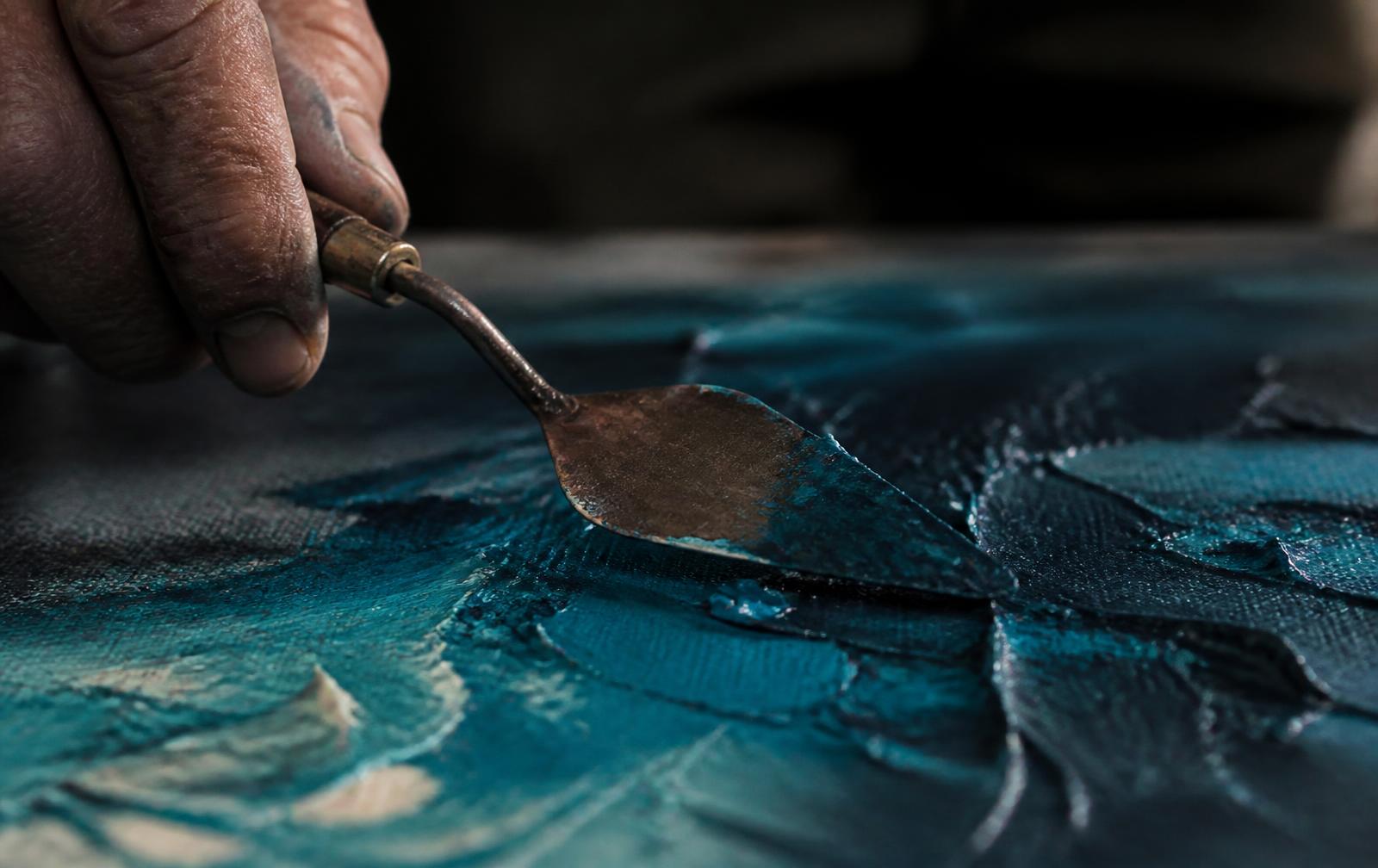 A copper palette knife pressing thick teal paint across a canvas